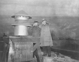 Photo of two men standing next to an air raid siren with hands over their ears.