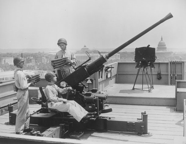 Photo of three soldiers manning an anti-aircraft gun on the top of a DC building, with the Capitol Building in the background.