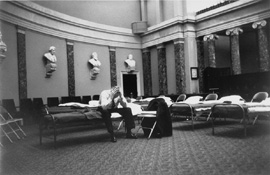 A man sits on a cot set up in the ornate Old Senate Chamber.